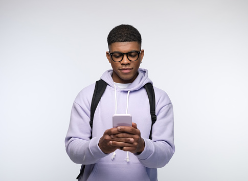 Confident african young man wearing lilac hoodie and backpack using smart phone. Studio portrait on white background.