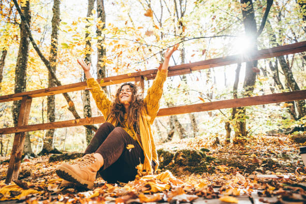 femme aux cheveux bouclés positifs en pull jaune jetant des feuilles sèches dans la forêt d’automne pittoresque avec des arbres colorés par temps ensoleillé. - nature forest clothing smiling photos et images de collection