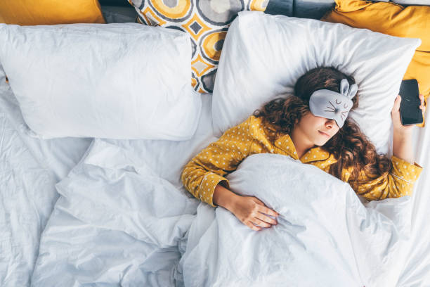 mujer millennial feliz acostada en la cama con máscara para dormir y teléfono en la mano. - máscara para los ojos fotografías e imágenes de stock