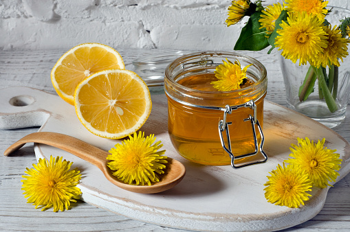 Honey from dandelions in glass jar, lemon slices and fresh flowers of dandelions on a white kitchen board. Rustic style. Close-up.