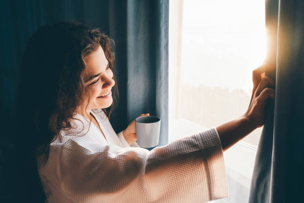 la signora dai capelli ricci in accappatoio bianco apre le tende per guardare fuori dalla grande finestra bevendo caffè caldo nella camera d'albergo buia all'alba. - waking up window women morning foto e immagini stock