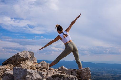 The healthy mid-adult woman at the top of the rocky mountain, during a fresh summer day, doing warrior 2 yoga pose