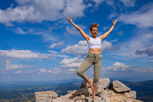 Mid-adult woman, doing an eagle yoga pose, while maintaining balance on the rocky top of the mountain