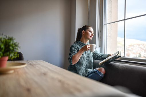 Beautiful Female Looking Out Window While Enjoying Coffee And Good Book