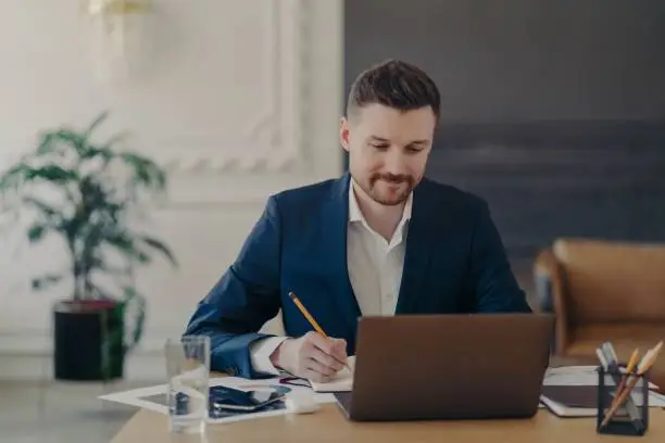 Photo of Indoor shot of handsome male office worker rewrites information from notepad in diary sits at desk dressed in formal clothes spends time for searching info. Businessman takes notes does paper work