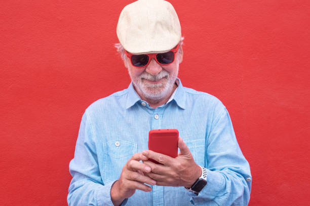 istock Senior bearded man standing on red background wearing cap and red sunglasses using mobile phone. Caucasian people smiling 1327810899