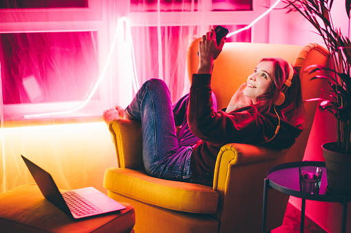 Young woman making video call with smart phone in living room with neon light at home .