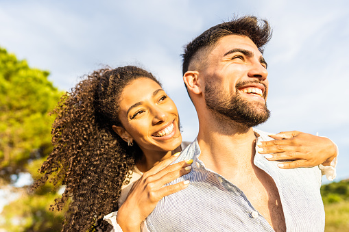 Multiracial happy young beautiful couple in love having fun outdoor in nature at dusk looking at horizon kissed by the setting sun. Toothy smiling Afro-American girl embracing from back her boyfriend