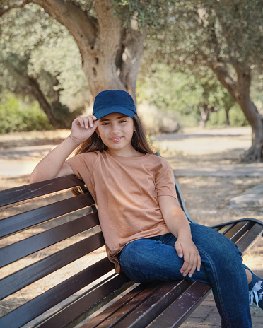 Smiling girl kid wearing t-shirt; jeans and baseball cap sits on the bench in the park