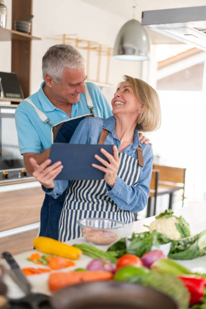 glückliches seniorenpaar, das zu hause nach einem rezept auf einem tablet kocht - senior adult digital tablet domestic life learning stock-fotos und bilder