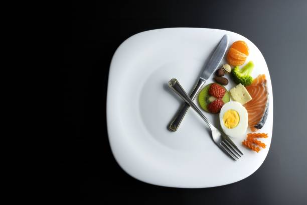 un plato simulando un reloj con las manecillas de un tenedor y un cuchillo que muestra la comida, concepto de ayuno intermitente - ayuno intermitente fotografías e imágenes de stock