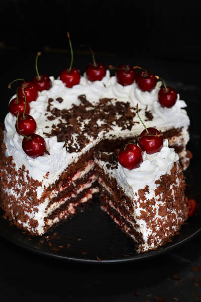 istock Close-up image of sliced Black Forest gateau with piped whipped cream rosettes with morello cherries, luxury chocolate cake frosted with whipped cream and covered with chocolate shavings, black plate, black background, focus on foreground, elevated view 1327824415