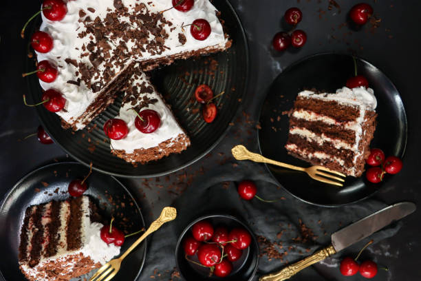 Image of sliced Black Forest gateau with piped whipped cream rosettes with morello cherries, luxury chocolate cake frosted with whipped cream and covered with chocolate shavings, black plate, black background, elevated view