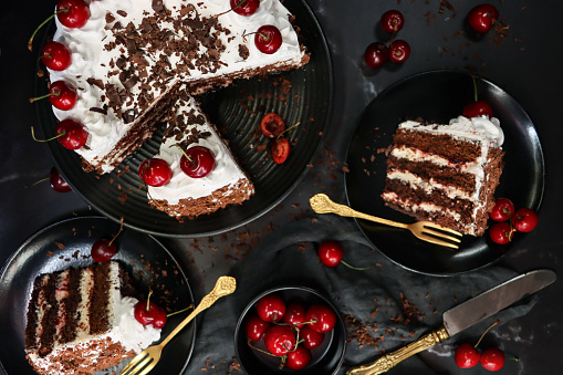 Stock photo showing an elevated view of plates containing sliced, homemade, luxury, Black Forest gateau. Served on black plates on a black surface surrounded by gold cutlery besides a bowl of morello cherries.