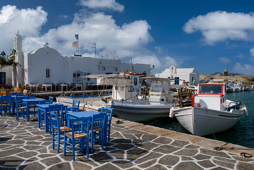 Blue tables and chairs of a quay side restaurant at the harbor of Naoussa in Greece. A very traditional greek scene with fishing boats an a small white church. Naoussa is a small town on Paros, one of the Cyclades Islands in Greece.