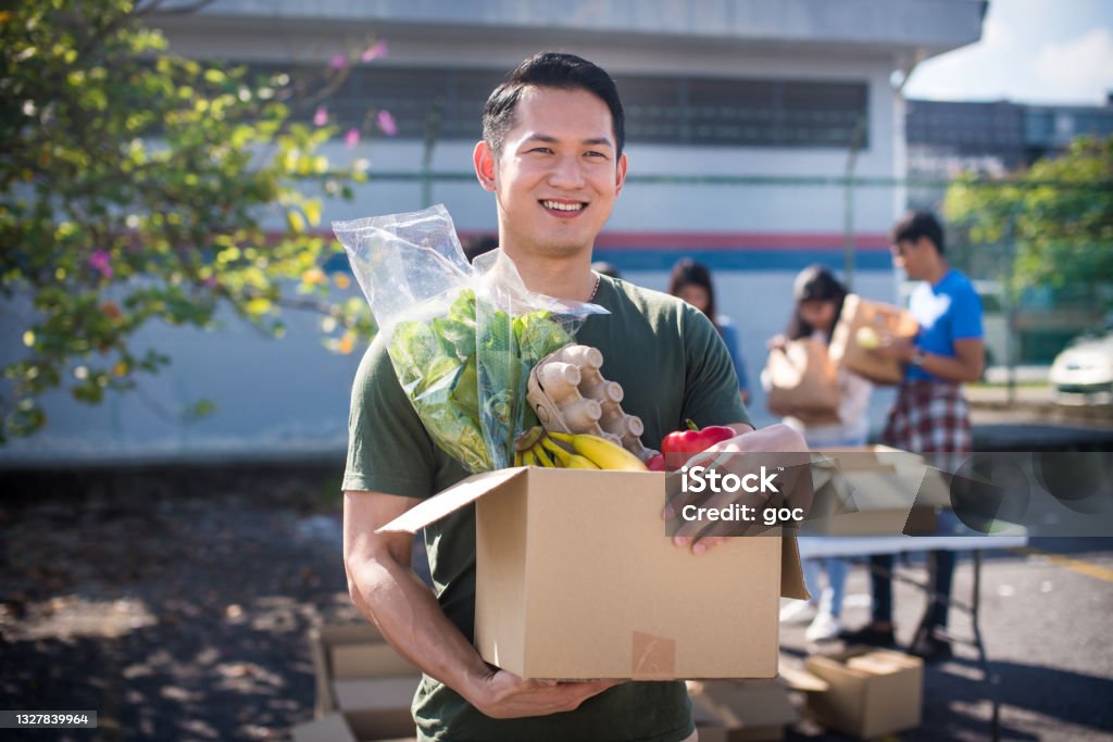 Cheerful and smiling Asian male volunteer and his colleagues distributing grocery food at community food bank A group of young Asian volunteers preparing grocery food into boxes for those needy people having financial difficulty during global economy crisis. Food box filled with fresh vegetables, fruits, cereals, milk, eggs and canned goods are deliver to the local needy community during a outdoor food drive. Volunteer Stock Photo