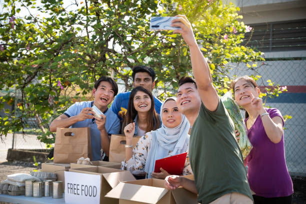 Asian volunteers taking group selfie after completing the task of give out free grocery food at charity food bank A group of young Asian volunteers enjoy taking selfie after successful distributing grocery food at community food bank. non profit organization photos stock pictures, royalty-free photos & images