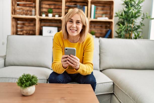 istock Middle age blonde woman using smartphone sitting on the sofa at home. 1327846318