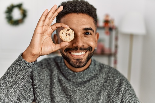 Young african american man smiling happy holding cookie over eye sitting on the table at home.