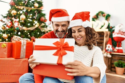 Middle age hispanic couple wearing christmas hat. Sitting on the floor opening gift at home.