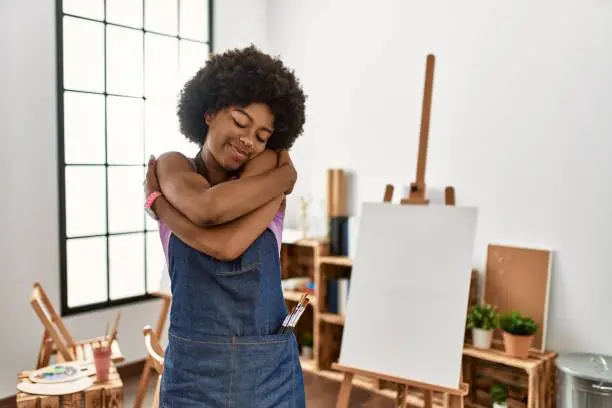 Young african american woman with afro hair at art studio hugging oneself happy and positive, smiling confident. self love and self care