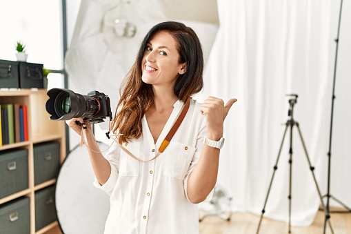 Beautiful caucasian woman working as photographer at photography studio smiling with happy face looking and pointing to the side with thumb up.