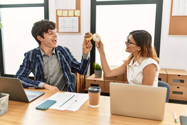Two business workers smiling happy toasting with doughnuts at the office.