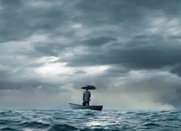 A man holding an umbrella stands with his briefcase on a boat that is stranded in the middle of the ocean.