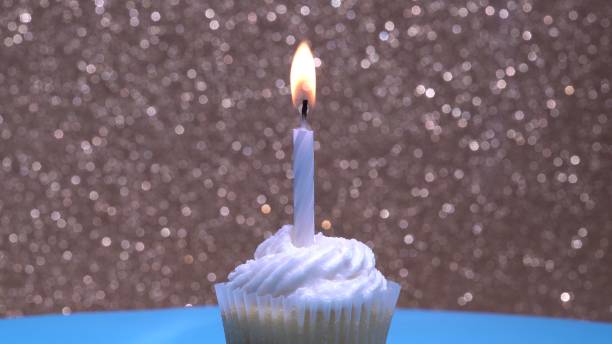 istock Cupcake with single birthday candle burning and sugar sprinkles icing on gold background. Delicious muffin decoration. Homemade vanilla cup cake with buttercream frosting. Shallow depth of field. 1327873551