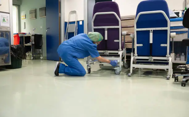Woman from the cleaning service of a hospital crouching cleaning the wheels of a patient chair in the operating room beforehand. Sanitary concept