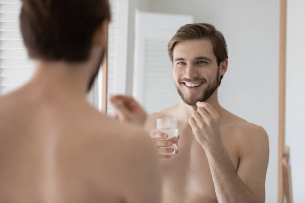Happy handsome shirtless guy taking vitamins, food supplement Happy handsome shirtless guy taking vitamins, food supplement, antidepressant, remedy, probiotics in bathroom, holding pill, drinking water, looking in mirror, smiling. Healthcare, pharmacy concept metrosexual stock pictures, royalty-free photos & images