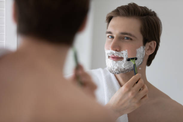 Positive attractive young guy with gel foam on lower face Positive attractive young guy with gel foam on lower face shaving with razor at mirror. Handsome man enjoying morning bath routine, caring for appearance, healthy facial skin. Hygiene, skincare concept shaving stock pictures, royalty-free photos & images