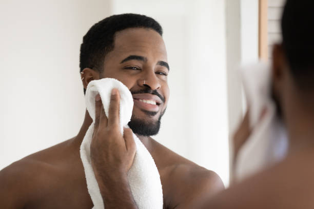 Happy handsome African American guy drying face with white towel Happy handsome African American guy drying face with white towel in bathroom, looking at mirror. Young man with bare muscular shoulders and stylish beard enjoying bath routine. Grooming concept stubble male african ethnicity facial hair stock pictures, royalty-free photos & images