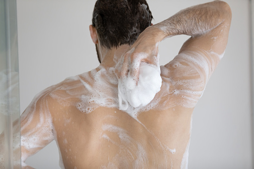 Back of handsome athletic wet young man taking shower, rubbing muscular body with foamy sponge, soap, cleansing gel. Male morning bath routine, daily hygiene, bodycare concept. Rear view