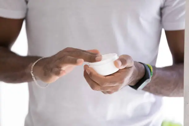 Hands of mixed raced Black man in white t-shirt, taking moisturizing collagen cream, lotion, mask, hydrating balm from jar. Male beauty care, skincare, cosmetic product concept. Close up, cropped shot