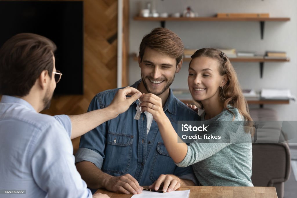 Happy young male Real Estate Agent giving keys to couple. Happy young male real estate agent giving keys from new apartment to sincere happy loving family couple. Smiling young husband and wife feeling excited of purchasing own dwelling, independence concept Real Estate Agent Stock Photo