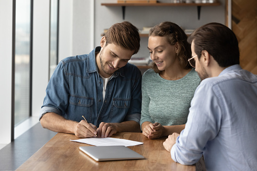 Happy young family couple clients signing insurance document, making good deal agreement with professional real estate agent, lawyer or broker at office meeting, celebrating purchasing own apartment.