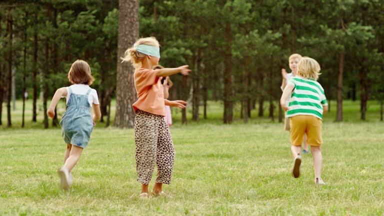 Group of kids playing blind man's buff tag game in park. Blindfolded little girl attempting to touch friend clapping hands and running around. Children having fun on summer vacation