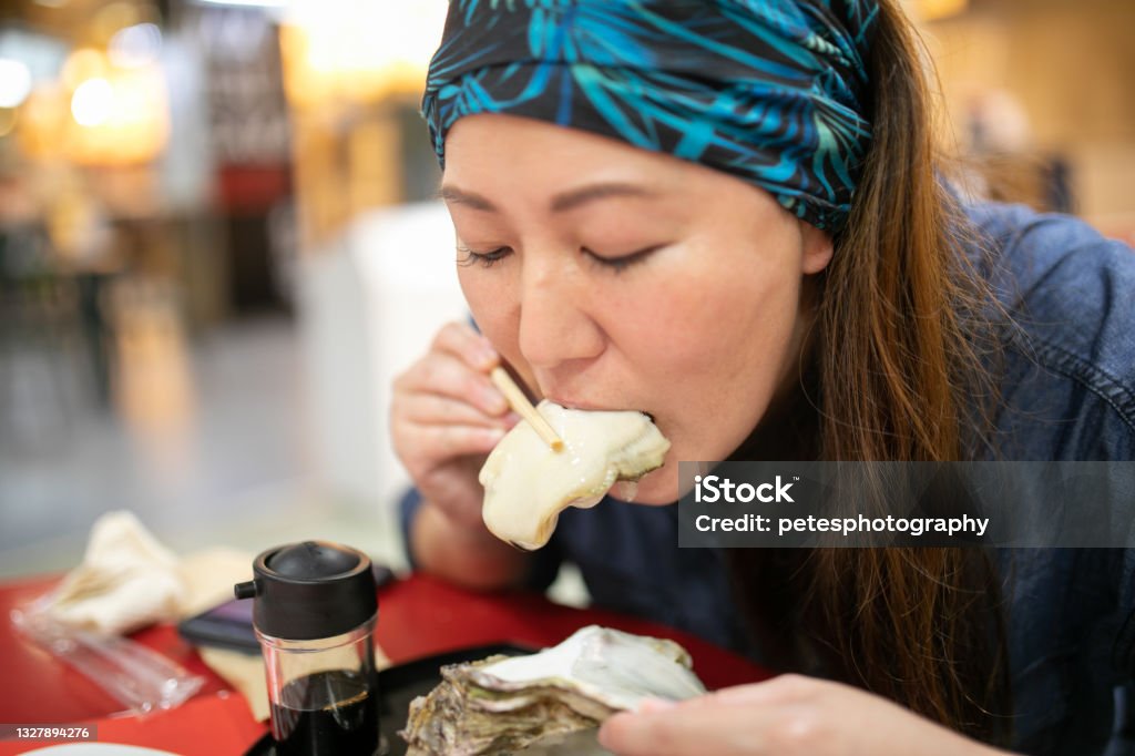 A woman eating a huge fresh Oyster from the shell A Japanese woman enjoying a massive size fresh Oyster with chopsticks from the shell indoors. 40-44 Years Stock Photo