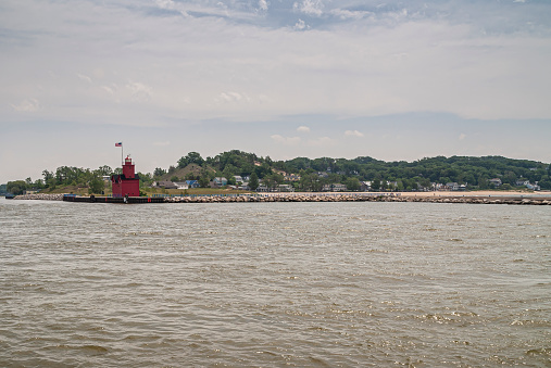 Holland, MI, USA - June 8, 2008: Wide shot from Michigan Lake of red harbor lighthouse at lands end under light blue sky. American flag on top. Forested dunes on horizon.