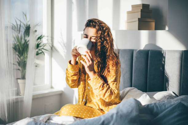 mulher de pijama amarelo sentada na cama e bebendo café no quarto da manhã ensolarada. - manhã - fotografias e filmes do acervo