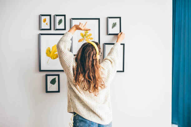 artiste femme expérimentée accroche des images d’appliques sur le mur avec des décorations écoutant de la musique dans des écouteurs dansant dans un nouvel appartement avec un fauteuil jaune. - armchair comfortable relaxation headphones photos et images de collection