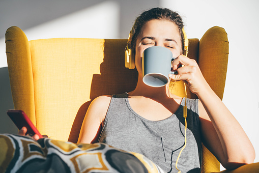 Relaxed young woman sitting on armchair listening to music and drinking coffee at home.