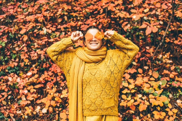 mujer joven juguetona con ropa naranja brillante con bufanda larga esconde ojos detrás de hojas de color naranja seco cerca de un gran arbusto rojo en el pintoresco bosque de otoño - autumn women leaf scarf fotografías e imágenes de stock