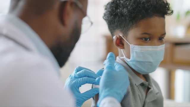 Male doctor of african american ethnicity or nurse giving shot or vaccine to a child age 9 yearold patient's shoulder at hospital or home to vaccination and prevention against coronavirus pandemic together.COVID-19 Vaccine concept.