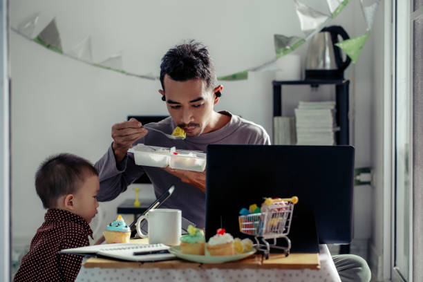 An Asian father and son are eating while looking at screen of electronic device of them at home. stock photo