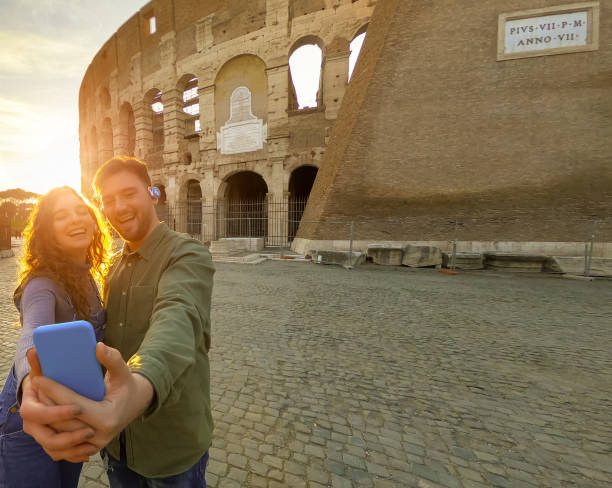 Young traveler couple using smartphone for videos, music and selfie. Happy tourists visiting Colosseum in sunset. Honeymoon trip, tourist, love holiday and vacation travel concept - Image pareja de jóvenes viajeros que utilizan teléfonos inteligentes para videos, música y selfie. turistas felices visitando el coliseo al atardecer. viaje de luna de miel, turista, vacaciones de amor y concepto de viaje de vacaciones - imagen