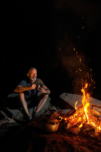 Relaxed mature man, tourist sitting near campfire, drinking coffee or tea while cooking dinner outdoors in forest at night. Camping, tourism concept