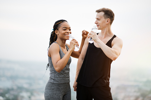 African woman and caucasian man in sport clothes standing outdoors, holding bottle of water and laughing to each other. Happy couple enjoying active workout on fresh air.