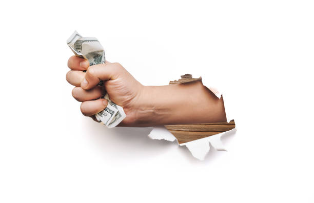 A male hand appears in the hole in torn white paper and squeezes two hundred-dollar bills. The concept of poverty, benefits, scholarships, and stinginess. stock photo